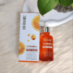 Dr. Rashel Vitamin C face serum in an orange glass dropper bottle placed next to its white box with orange slice graphics, photographed on a soft cream background with green leaves, highlighting a brightening and anti-aging formula with hyaluronic acid.