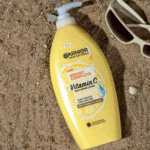 Yellow Garnier Skin Naturals Bright Complete Vitamin C body serum lotion with white pump placed on sandy beach surface, with sunglasses and seashells nearby, highlighting lemon and vitamin C graphics on the bottle.