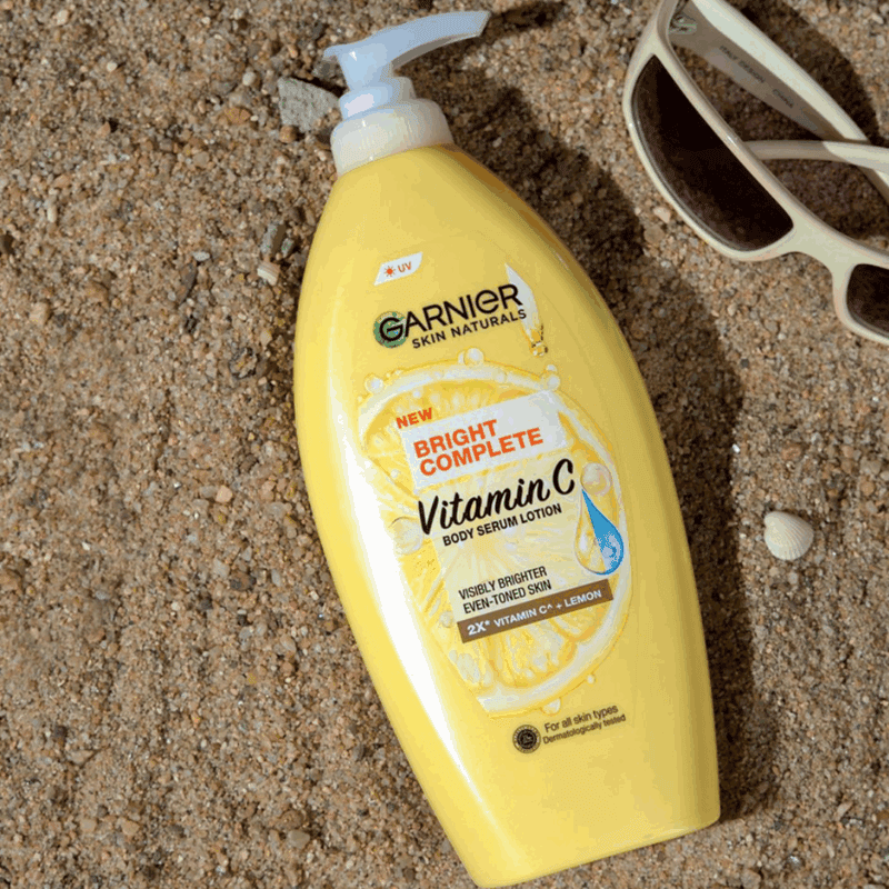 Yellow Garnier Skin Naturals Bright Complete Vitamin C body serum lotion with white pump placed on sandy beach surface, with sunglasses and seashells nearby, highlighting lemon and vitamin C graphics on the bottle.
