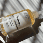 The Ordinary Glycolic Acid 7% Toning Solution bottle lying on its side in natural sunlight, showing the clear bottle, white label, nozzle cap, and light amber liquid with soft shadow patterns.
