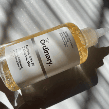 The Ordinary Glycolic Acid 7% Toning Solution bottle lying on its side in natural sunlight, showing the clear bottle, white label, nozzle cap, and light amber liquid with soft shadow patterns.