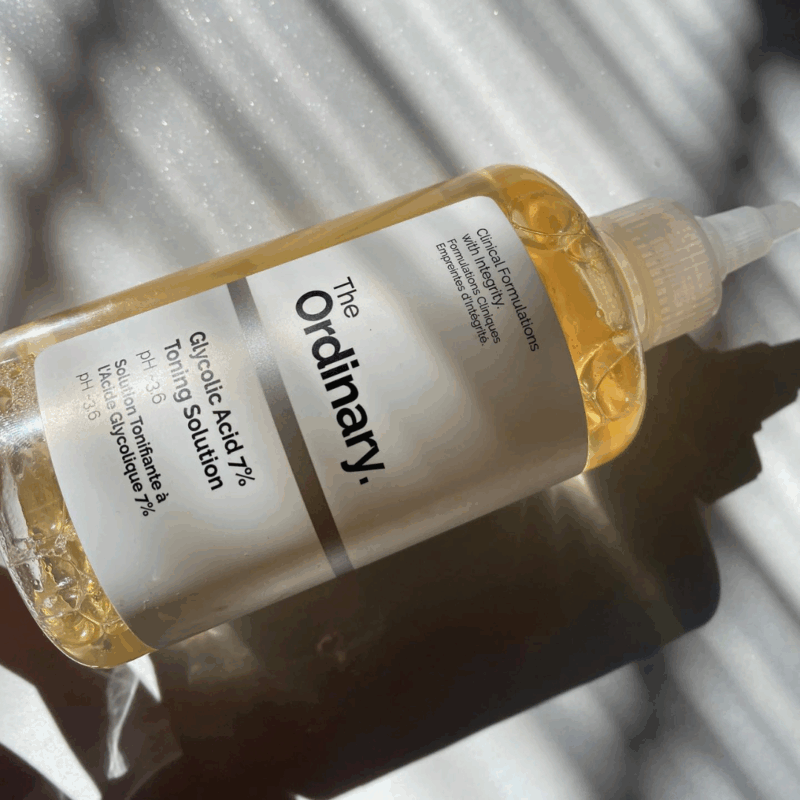 The Ordinary Glycolic Acid 7% Toning Solution bottle lying on its side in natural sunlight, showing the clear bottle, white label, nozzle cap, and light amber liquid with soft shadow patterns.