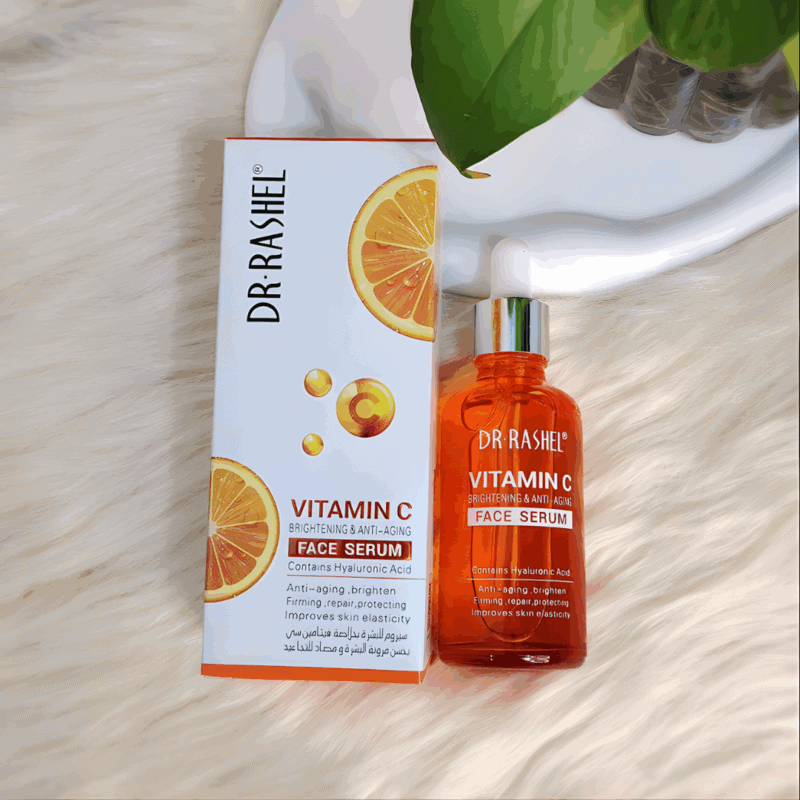 Dr. Rashel Vitamin C face serum in an orange glass dropper bottle placed next to its white box with orange slice graphics, photographed on a soft cream background with green leaves, highlighting a brightening and anti-aging formula with hyaluronic acid.