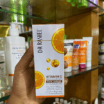 Hand holding Dr. Rashel Vitamin C face serum box featuring orange slice graphics and Vitamin C icons, displayed in a store shelf setting with skincare products in the background, highlighting a brightening and anti-aging formula with hyaluronic acid.
