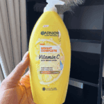 Hand holding a yellow Garnier Skin Naturals Bright Complete Vitamin C body serum lotion with white pump, photographed indoors near curtains and shelves, showing lemon graphic and “2X Vitamin C + Lemon” label on the bottle.