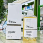 The Ordinary Glycolic Acid 7% Toning Solution bottle placed beside its original white box on a store counter, showing the clear bottle, white label, light amber liquid, and blurred skincare shelves in the background.