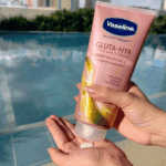 Person squeezing Vaseline Healthy Bright Gluta-Hya Dewy Radiance serum burst lotion onto their palm by a swimming pool, showing the pink 330ml tube with GlutaGlow, Hyaluron, and Niacinamide branding.