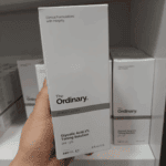Hand holding The Ordinary Glycolic Acid 7% Toning Solution box on a white retail shelf, showing the minimalist grey packaging, product name, pH 3.6 details, and similar boxes stacked in the background.
