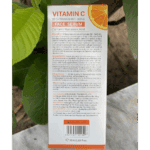 Back view of Dr. Rashel Vitamin C Brightening & Anti-Aging Face Serum box showing ingredients, benefits, usage directions, warnings, and manufacturer details, photographed outdoors with green leaves and natural light background.