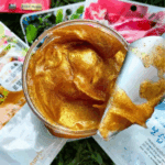 Top-view close-up of La’Fresh Gold exfoliating face and body scrub with a rich, shimmering gold texture inside an open jar, shown with a spatula lifting the scrub, surrounded by skincare sheet masks in the background.
