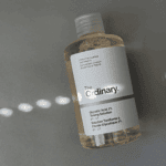 The Ordinary Glycolic Acid 7% Toning Solution bottle placed on a grey surface, photographed from above with soft natural light and shadow patterns, showing the clear liquid, minimalist white label, and pH 3.6 details.