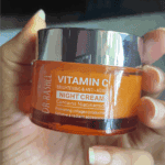 Close-up of Dr. Rashel Vitamin C Brightening Night Cream jar held in hand, featuring an orange translucent container with a silver lid, highlighting its anti-aging formula with vitamin C and niacinamide.