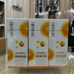 Three Dr. Rashel Vitamin C face serum boxes standing side by side on a shop counter, featuring orange slice graphics and Vitamin C icons, highlighting a brightening and anti-aging formula with hyaluronic acid in a retail store setting.