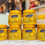 Front view of multiple La’Fresh Gold deep exfoliating face and body scrub jars stacked and arranged on a store counter, featuring bright gold scrub texture inside clear containers with yellow lids and blurred retail shelves in the background.