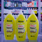 Three yellow Garnier Skin Naturals Bright Complete Vitamin C body serum lotion bottles displayed side by side on a glass shelf, with a cosmetics store background showing various skincare products on shelves behind.