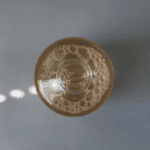 Top-down view of The Ordinary Glycolic Acid 7% Toning Solution bottle showing the clear golden liquid with visible air bubbles inside, placed on a grey surface with soft natural light reflections.