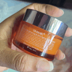 Back view of Dr. Rashel Vitamin C Brightening & Anti-Aging Night Cream jar held in hand, showing the orange translucent container with a reflective silver lid and printed product details on the label.