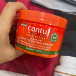 Close-up of Cantu Shea Butter Leave-In Conditioning Cream jar held in hand on fabric background