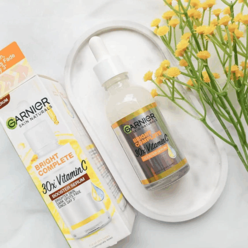 Garnier Bright Complete 30x Vitamin C Booster Serum bottle displayed with original packaging on a white background with flowers