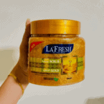Hand holding a La’Fresh Gold face and body scrub jar with a yellow lid, showing the gold-textured exfoliating scrub inside a clear container against a plain background.
