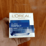 Hand holding L’Oréal Paris Aura Perfect Night Cream box showing fights dark spots and evens skin tone message