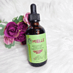 Mielle Rosemary Mint Scalp and Hair Strengthening Oil bottle placed on white fur background with purple roses