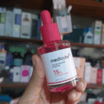 Hand holding Medicube TXA Niacinamide 15 Serum 30ml pink dropper bottle with skincare shelves in background