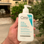 A hand holding a bottle of CeraVe Acne Control Cleanser outdoors, with a blurred house and greenery in the background, showcasing the white and teal packaging designed to treat acne and control excess oil.