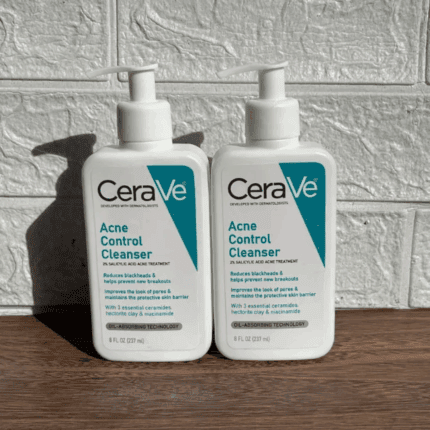 Two bottles of CeraVe Acne Control Cleanser placed side by side on a wooden surface against a textured wall, featuring white and teal packaging with pump dispensers for acne treatment and oil control.
