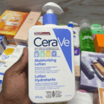 A hand holding a bottle of CeraVe Baby Moisturizing Lotion 473ml with white and blue packaging, surrounded by various skincare products, designed to hydrate and protect delicate baby skin.