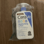 A sealed value-size bottle of CeraVe Baby Moisturizing Lotion 473ml wrapped in plastic, placed on a wooden surface, designed to hydrate and protect baby’s delicate skin with ceramides.