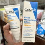 A hand holding a CeraVe Facial Moisturising Lotion AM SPF 50 box alongside the pump tube, showing product details for normal to dry skin with SPF 50 UV protection against a warm indoor background.
