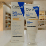 A CeraVe Facial Moisturising Lotion AM SPF 50 shown with its packaging box and pump tube, placed on a countertop with a softly blurred indoor background, highlighting features for normal to dry skin and broad-spectrum UV protection.