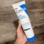 A hand holding a tube of CeraVe Gentle Hydrating Conditioner with water droplets on the packaging, shown against a wooden floor background, highlighting its gentle, hydrating formula for hair and scalp care.