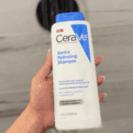 A hand holding a bottle of CeraVe Gentle Hydrating Shampoo 355ml under running water in a shower, with a showerhead in the background, highlighting its gentle cleansing and hydrating formula.