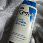 A bottle of CeraVe Gentle Hydrating Shampoo 355ml placed on a silky satin surface with soft lighting and a decorative feather, highlighting its gentle cleansing formula with ceramides and hyaluronic acid.