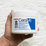 A hand holding a jar of CeraVe Moisturizing Cream against a textured white background, showing the product’s label with blue branding, fragrance-free formula, and benefits for dry to very dry skin.