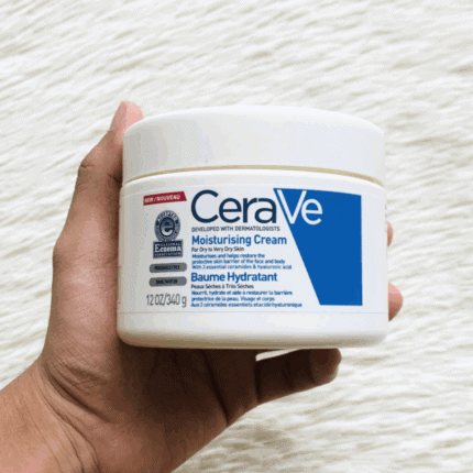 A hand holding a jar of CeraVe Moisturizing Cream against a textured white background, showing the product’s label with blue branding, fragrance-free formula, and benefits for dry to very dry skin.