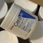 A close-up of a CeraVe Moisturizing Cream jar placed among several similar white containers, showing the product’s blue and white label designed for dry to very dry skin with ceramides and hyaluronic acid.
