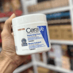 A hand holding a jar of CeraVe Moisturizing Cream for dry to very dry skin, with a blurred background of shelves, highlighting the product’s white packaging with blue branding and eczema association seal.