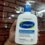 Hand holding Cetaphil Daily Facial Cleanser 473ml bottle for combination to oily sensitive skin, fragrance free face wash with hydrating glycerin and vitamins B5 and B3, store background.