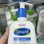 Close-up of a hand holding Cetaphil Oily Skin Cleanser 236ml bottle with pump, showing label details for oily and sensitive skin, displayed in a retail shelf setting.