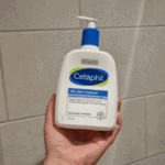 Hand holding a bottle of Cetaphil Oily Skin Cleanser with pump dispenser against a tiled background, showing real-life size and packaging for oily and sensitive skin.