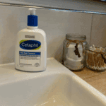 Cetaphil Oily Skin Cleanser bottle placed on a bathroom sink beside jars of cotton pads and earbuds, showing a real-life skincare setup for oily and sensitive skin.
