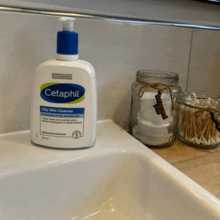 Cetaphil Oily Skin Cleanser bottle placed on a bathroom sink beside jars of cotton pads and earbuds, showing a real-life skincare setup for oily and sensitive skin.
