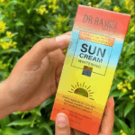 Hand holding Dr Rashel Sun Cream Whitening SPF 75 60g sunscreen with long lasting UVA UVB protection and oil control, displayed outdoors with green plants background.
