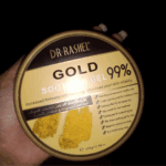A hand holding a jar of Dr. Rashel Gold Soothing Gel 99% (300g) with a gold lid, showing product claims for skin vitality, hydration, and soothing benefits against a dark background.