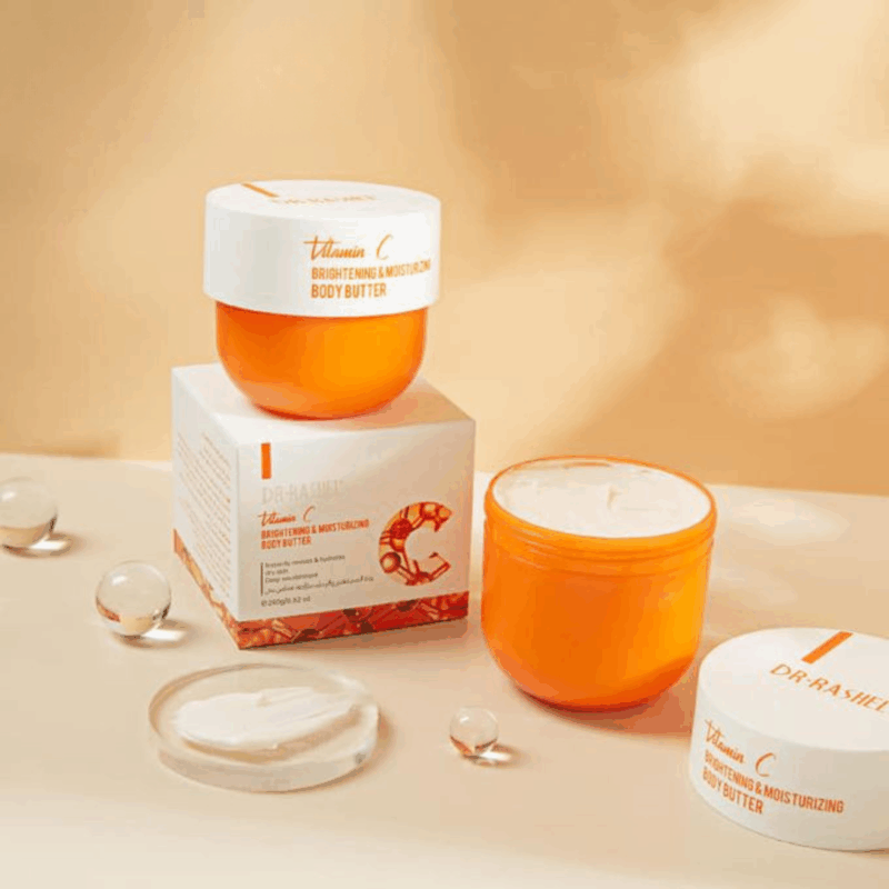 Multiple jars of Dr. Rashel Vitamin C Brightening and Moisturizing Body Butter displayed with packaging, including an open jar showing the cream texture, arranged with decorative elements on a warm-toned background.