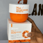 A hand holding a jar of Dr. Rashel Vitamin C Brightening and Moisturizing Body Butter placed on top of its packaging box, highlighting its hydrating and skin-brightening benefits against an indoor background.