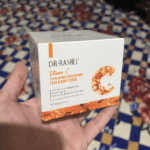 A hand holding the box of Dr. Rashel Vitamin C Exfoliating and Brightening Face and Body Scrub (250g), shown against a patterned indoor background.